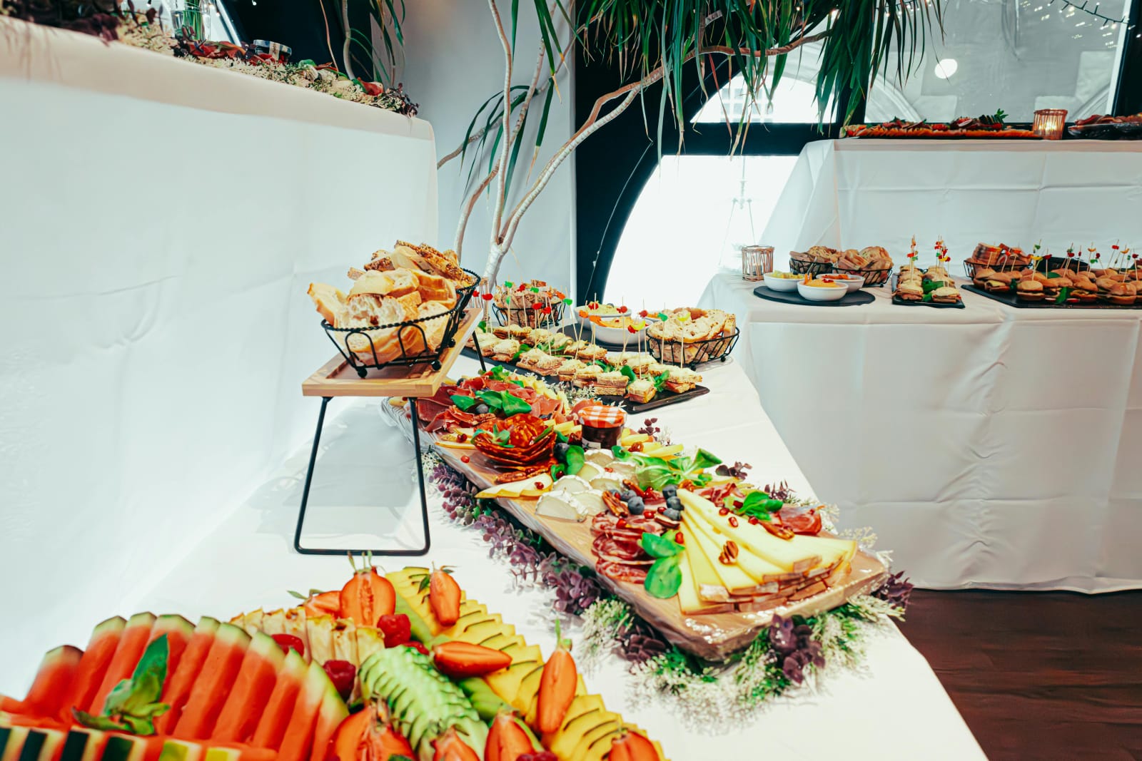 A long party buffet table with white linens featuring multiple levels of food including fruit platters, charcuterie boards, and bread baskets on wire risers.